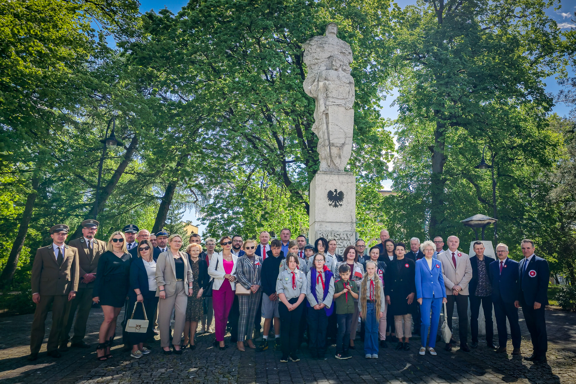 Uczestnicy uroczystości patriotycznej zgromadzeni przed pomnikiem wdzięczności w Łobzie, wśród nich władze gminne i powiatowe, służby mundurowe, przedstawieciele jednostek oświatowych i harcerze, otoczeni zielenią parku.