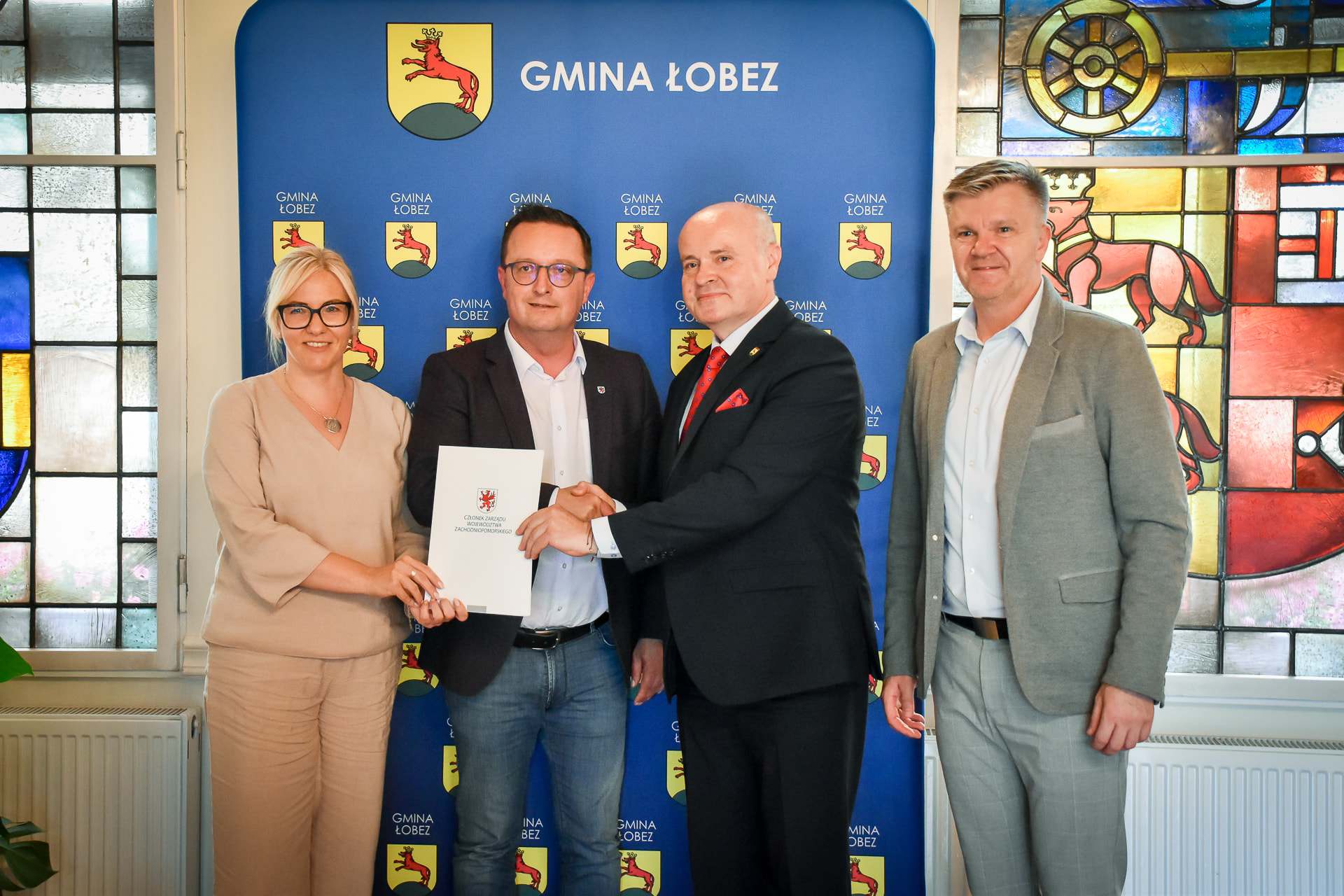 Uroczyste podpisanie dokumentu w Urzędzie Miejskim w Łobzie – cztery osoby stoją na tle ścianki z herbem Gminy Łobez; dwóch mężczyzn uściskiem dłoni symbolicznie przypieczętowuje współpracę, jedna z osób trzyma teczkę z logo Zachodniopomorskiego Urzędu Marszałkowskiego.