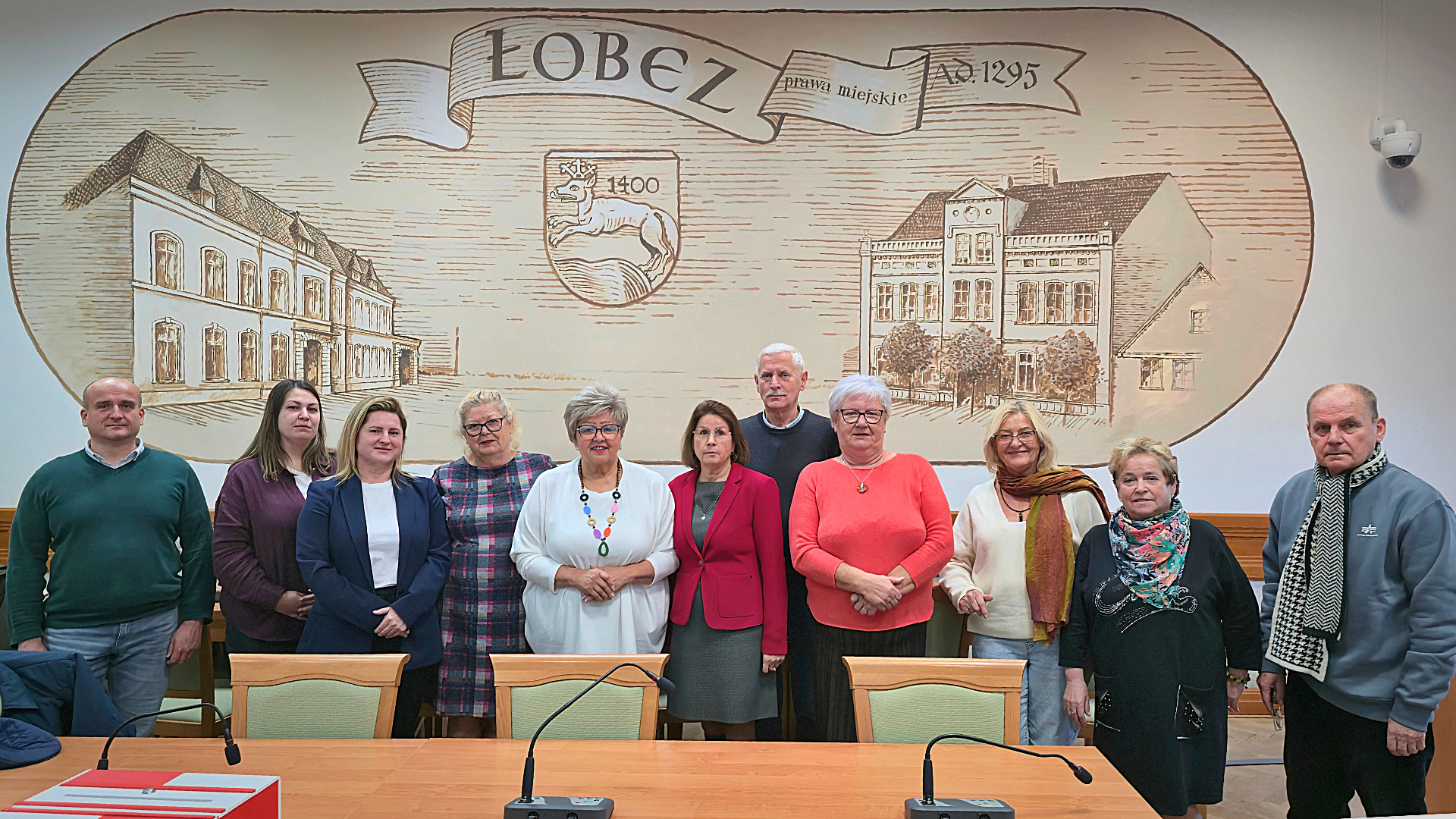Grupa ludzi pozuje do wsp&oacute;lnego zdjęcia w&nbsp;sali sesyjnej urzedu miejskiego w&nbsp;łobzie. W tle na&nbsp;ścianie rysunek przedstawiający stare budynki w&nbsp;Łobzie oraz&nbsp;datę nadania praw miejskich. 