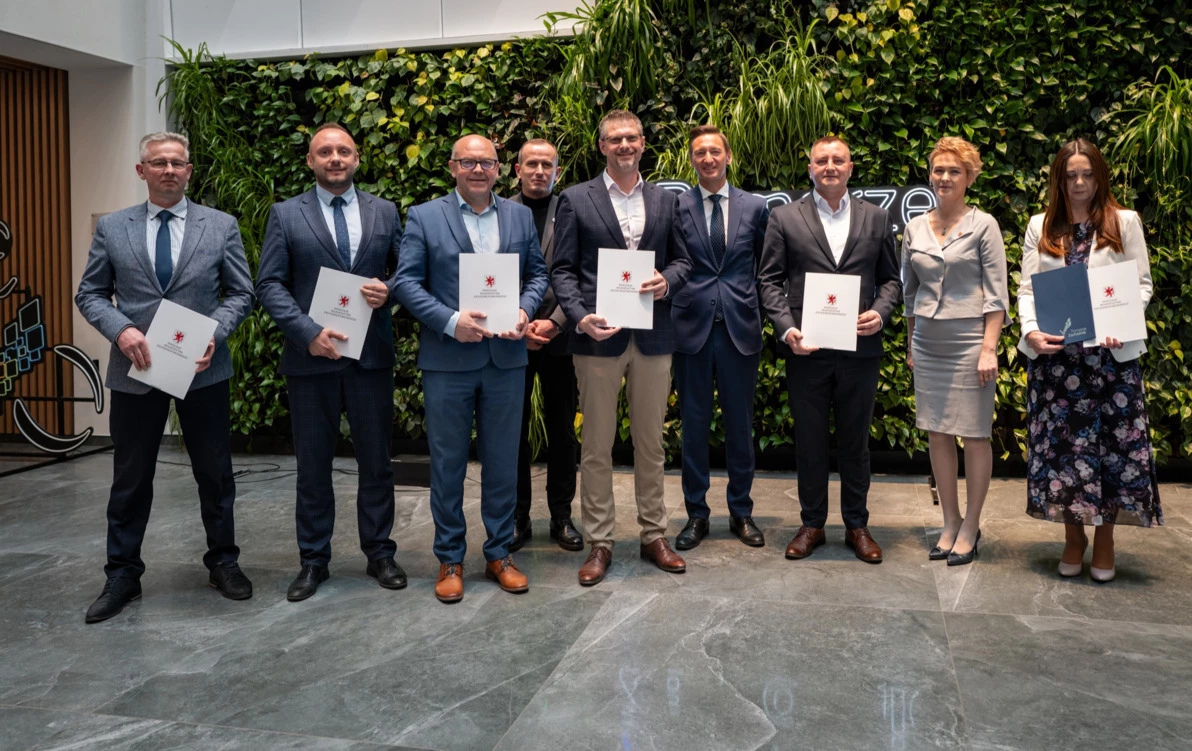 Grupowe zdjęcie przedstawiające dziewięć osób – kobiety i mężczyzn – stojących na tle zielonej ściany z roślinnością. Osoby w eleganckich strojach trzymają w dłoniach teczki z herbem Województwa Zachodniopomorskiego.